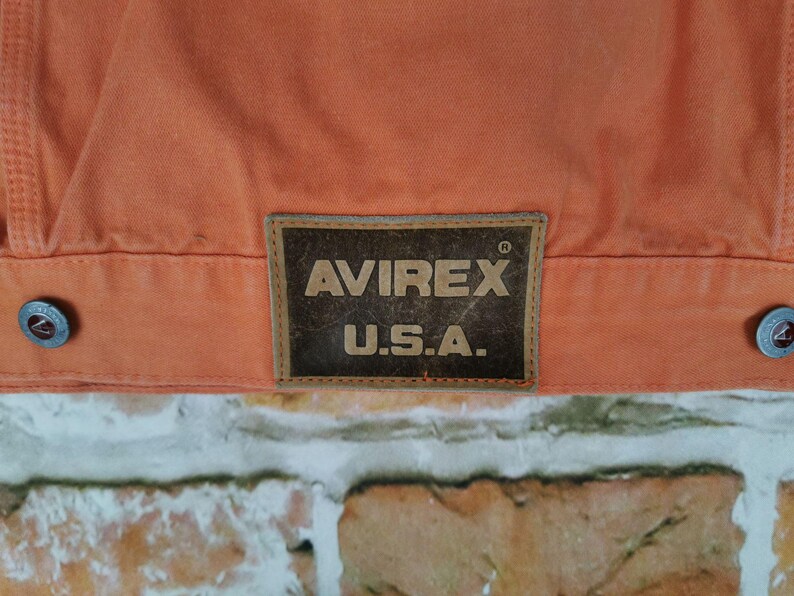 Puede incluir: Primer plano de una chaqueta naranja con el parche del logotipo "AVIREX U.S.A.". La chaqueta tiene dos botones plateados y una costura. El fondo es una pared de ladrillos.