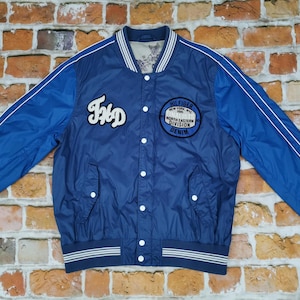 Op de afbeelding: Een blauw en wit baseballjack met een witte streep op elke mouw. Het jack heeft een knoopsluiting en een patch op de linkerborst met de tekst "Hilfiger New York 1985 North Eastern Division Denim". Het jack heeft ook een patch aan de linkerkant met de tekst "FHD".