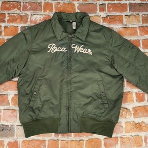 Könnte beinhalten: Eine dunkelgrüne Bomberjacke mit einem weißen gestickten Logo, auf dem "Roca Wear" steht.