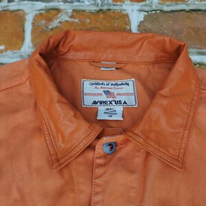 Puede incluir: Primer plano de una camisa abotonada naranja con cuello de cuero. La camisa tiene una etiqueta que dice "AVIREX U.S.A." y "Certificate of Authenticity". La camisa es talla XL.