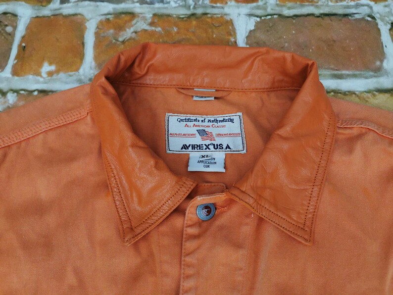Puede incluir: Primer plano de una camisa abotonada naranja con cuello de cuero. La camisa tiene una etiqueta que dice "AVIREX U.S.A." y "Certificate of Authenticity". La camisa es talla XL.