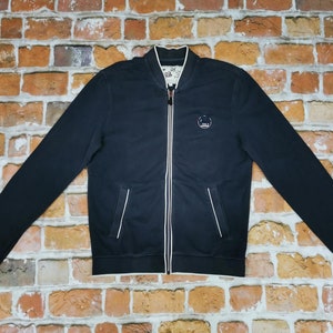 Puede incluir: Una chaqueta bomber azul marino con cremallera blanca y ribetes blancos. La chaqueta tiene un pequeño logotipo bordado en el pecho izquierdo.