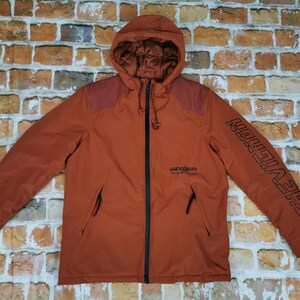 Op de afbeelding: Een roestkleurige capuchontrui met een ritssluiting en een zwarte rits. De trui heeft een patch op de linker schouder en de tekst "CHEVRON" op de linker borst. De rechter mouw heeft de tekst "LIFE REVOLUTION" verticaal bedrukt.