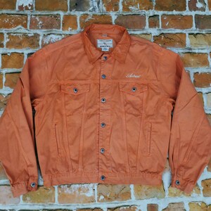 Puede incluir: Chaqueta vaquera naranja con botones, bolsillos en el pecho y cuello. La palabra "Astral" est&aacute; bordada en el pecho. La chaqueta se muestra sobre un fondo de pared de ladrillo.