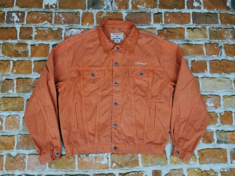 Puede incluir: Chaqueta vaquera naranja con botones, bolsillos en el pecho y cuello. La palabra "Astral" est&aacute; bordada en el pecho. La chaqueta se muestra sobre un fondo de pared de ladrillo.