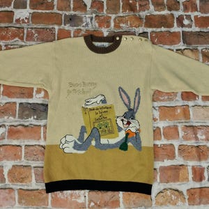 Könnte beinhalten: Ein beigefarbener Langarmpullover mit braunem Kragen und grünen Bündchen. Er zeigt eine Grafik von Bugs Bunny, der ein gelbes Buch mit dem Titel Jean de la Fontaine le lièvre et la tortue liest, mit dem Text Bugs Bunny Go To School darüber.