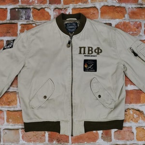 Può includere: Una giacca bomber beige con chiusura a zip e collo e polsini a costine marroni. La giacca ha una toppa sul petto sinistro con il testo "PBF" e "INFANTRY UNIVERSITY".