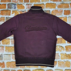 Puede incluir: Una sudadera con cremallera de color burdeos con cuello y puños de canalé. La sudadera tiene un gran logotipo "Champion" bordado en la espalda.