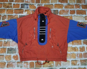 Puede incluir: Una chaqueta de esquí roja, azul y negra con un diseño de estrella en la parte delantera. La chaqueta tiene dos bolsillos con cremallera en la parte delantera y un cordón en la parte inferior.