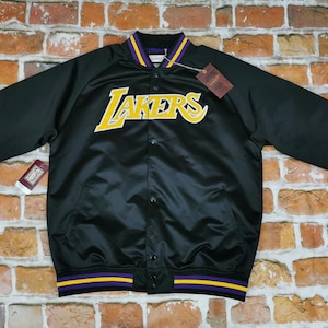 Peut inclure: Veste bomber en satin noir avec un col et des poignets rayés violet et jaune. La veste présente un grand logo jaune "LAKERS" sur le devant.