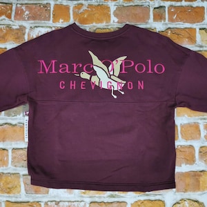 Puede incluir: Una sudadera burdeos con mangas largas. La parte trasera presenta el texto "Marc O'Polo CHEVIGNON" en rosa, junto con un gráfico de un pájaro volando. La sudadera se muestra sobre un fondo de pared de ladrillo.