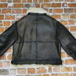 Pu&ograve; includere: Giacca in pelle marrone con colletto e polsini in montone color crema. La giacca presenta cuciture blu scuro e un classico stile aviatore. Il retro della giacca &egrave; visibile, mostrando le cuciture e i pannelli.
