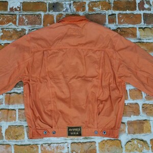 Puede incluir: Chaqueta naranja con cuello abotonado y mangas largas. La parte trasera de la chaqueta presenta un canes&uacute; y costuras verticales. Un peque&ntilde;o parche rectangular con "AVIREX U.S.A." est&aacute; cerca del dobladillo. Sobre un fondo de pared de ladrillos.