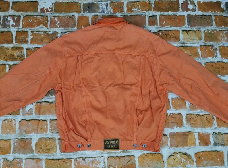 Puede incluir: Chaqueta naranja con cuello abotonado y mangas largas. La parte trasera de la chaqueta presenta un canes&uacute; y costuras verticales. Un peque&ntilde;o parche rectangular con "AVIREX U.S.A." est&aacute; cerca del dobladillo. Sobre un fondo de pared de ladrillos.