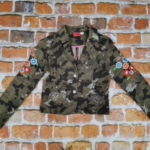 Könnte beinhalten: Eine Jacke im Tarnmuster mit Blumenstickerei an den Ärmeln. Die Jacke hat ein rosafarbenes Futter und zwei Knöpfe. Der Text "Girls Rule" ist auf der Vorderseite gestickt.