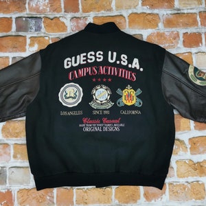 Op de afbeelding: Zwart varsity jack met leren mouwen. De achterkant heeft de tekst "GUESS U.S.A. CAMPUS ACTIVITIES" met logo's en de tekst "LOS ANGELES SINCE 1981 CALIFORNIA".