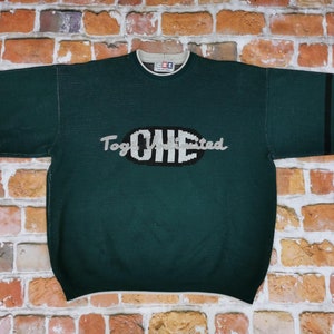 Puede incluir: Un suéter de punto verde oscuro con un logotipo blanco y negro que dice "Together We Cheered". El suéter tiene rayas negras en las mangas.