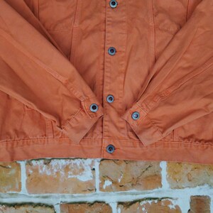 Puede incluir: Primer plano de una chaqueta vaquera naranja. La chaqueta tiene una parte delantera abotonada, dos bolsillos en el pecho y pu&ntilde;os abotonados. Las mangas est&aacute;n dobladas a la altura de los codos. La chaqueta est&aacute; hecha de un material vaquero resistente y es de color naranja liso.