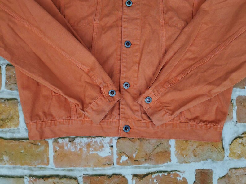Puede incluir: Primer plano de una chaqueta vaquera naranja. La chaqueta tiene una parte delantera abotonada, dos bolsillos en el pecho y pu&ntilde;os abotonados. Las mangas est&aacute;n dobladas a la altura de los codos. La chaqueta est&aacute; hecha de un material vaquero resistente y es de color naranja liso.