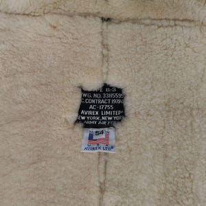 Pu&ograve; includere: Primo piano dell'interno di una giacca in montone. La fodera in lana beige &egrave; visibile, con un'etichetta nera con testo tra cui "AVIREX LIMITED" e "U.S. ARMY AIR FORCE". Un'etichetta pi&ugrave; piccola mostra "54 IN" e la bandiera americana.