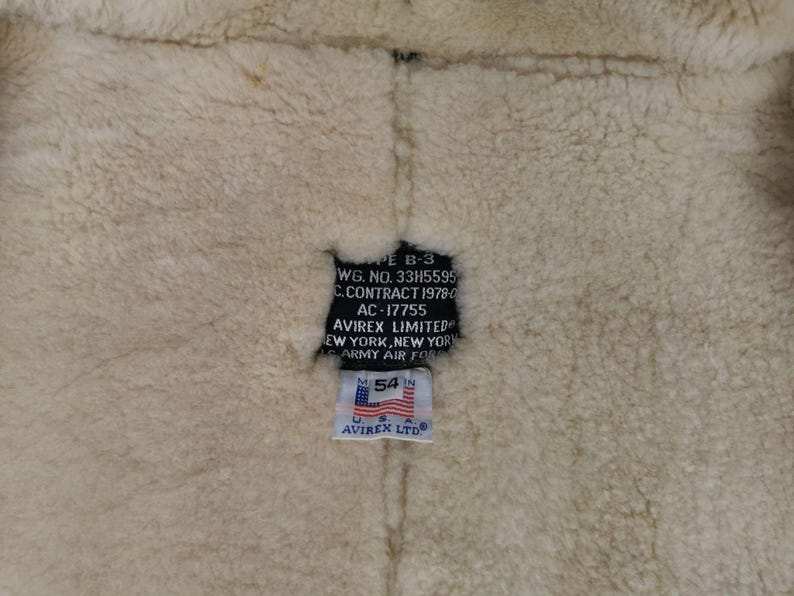 Pu&ograve; includere: Primo piano dell'interno di una giacca in montone. La fodera in lana beige &egrave; visibile, con un'etichetta nera con testo tra cui "AVIREX LIMITED" e "U.S. ARMY AIR FORCE". Un'etichetta pi&ugrave; piccola mostra "54 IN" e la bandiera americana.