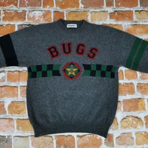 Könnte beinhalten: Grauer Pullover mit schwarzen und grünen Streifen und dem Wort "BUGS" in großen Buchstaben über der Brust. Der Pullover hat ein kariertes Muster und einen Stern mit einem Schlüssel in der Mitte.
