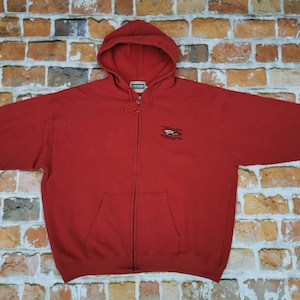 Puede incluir: Una sudadera con capucha roja con cremallera y un pequeño diseño bordado en la parte delantera. La sudadera tiene una capucha con cordón y un bolsillo canguro.