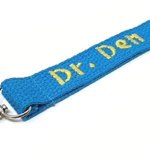 May include: A blue lanyard with a silver swivel hook and a gold embroidered name "Dr. Den".