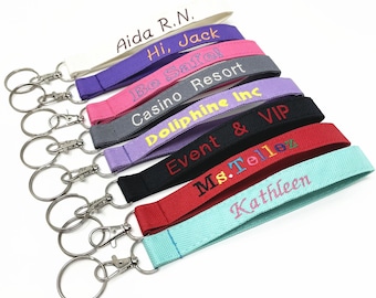 May include: A collection of embroidered wrist lanyards in various colors, including white, purple, pink, gray, lavender, black, red, and teal. Each lanyard features text in different colors and includes a silver-colored key ring.