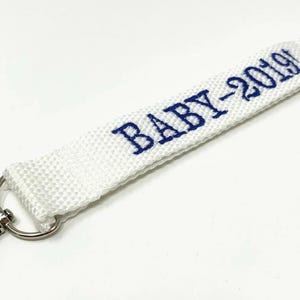 May include: White fabric keychain with a silver clip and a silver ring. The fabric has the text "BABY-2019!" embroidered in blue.