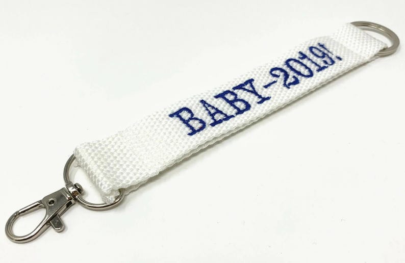 May include: White fabric keychain with a silver clip and a silver ring. The fabric has the text "BABY-2019!" embroidered in blue.