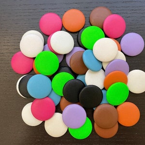 May include: A collection of colorful, round buttons in various shades, including pink, orange, green, blue, white, black, brown, and purple. The buttons are scattered on a dark surface, creating a vibrant and playful display.