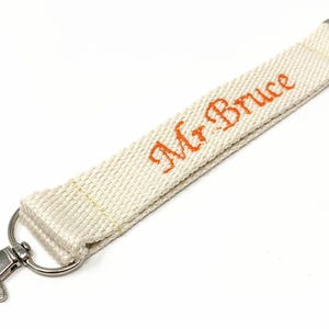 May include: A white fabric key chain with a silver swivel clasp and a silver ring. The fabric has the text "Mr. Bruce" embroidered in orange.