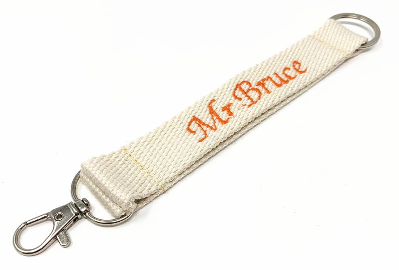 May include: A white fabric key chain with a silver swivel clasp and a silver ring. The fabric has the text "Mr. Bruce" embroidered in orange.