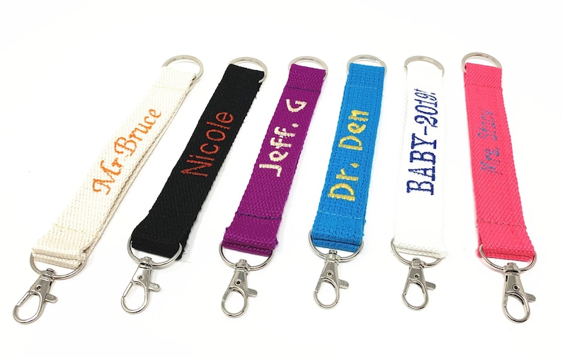 May include: Six colorful keychains with personalized embroidered text. The keychains are made of fabric and have silver metal key rings. The text on the keychains includes "Mr. Bruce", "Nicole", "Jeff. G", "Dr. Den", "BABY-2018", and "Mrs. Smith".