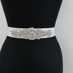 May include: A white satin bridal belt with a large, ornate rhinestone embellishment. The embellishment is made up of several clusters of clear rhinestones.