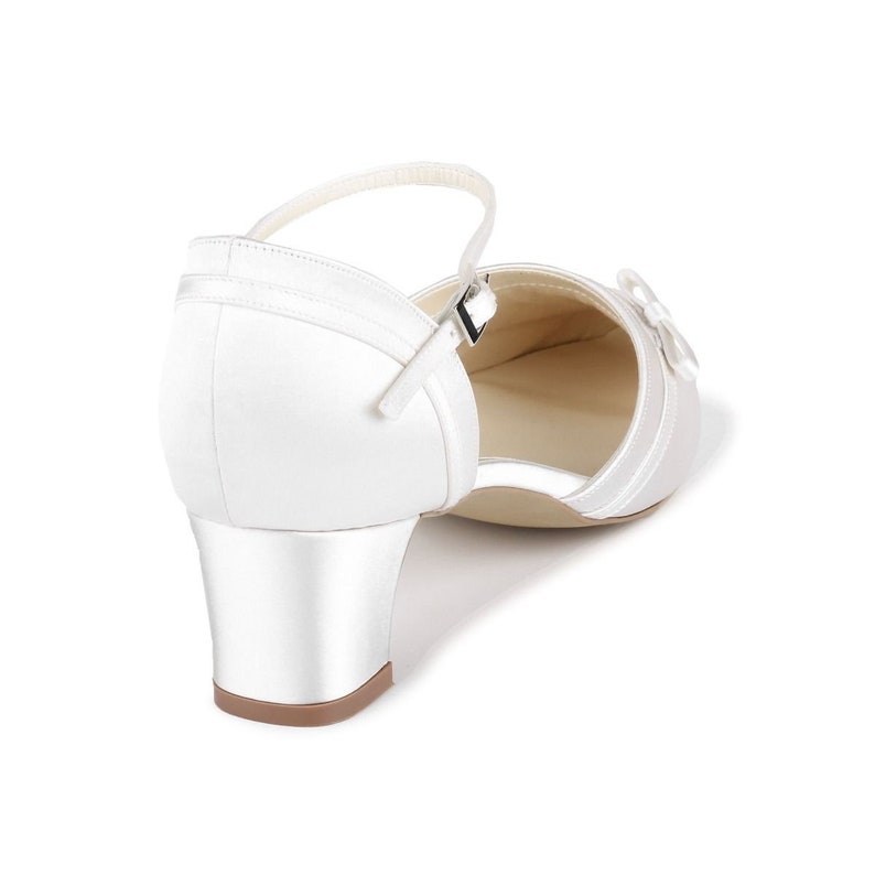 May include: White satin bridal shoes with a low block heel and an ankle strap. The shoes have a satin bow on the toe.