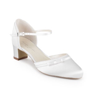 May include: White satin bridal shoes with a bow detail and an ankle strap with a buckle. The shoes have a block heel.