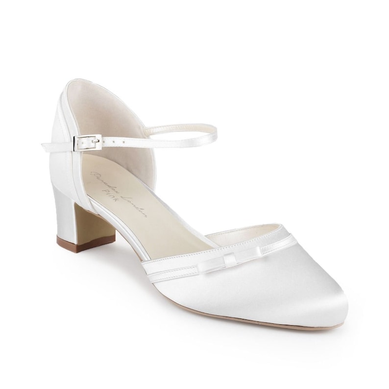 May include: White satin bridal shoes with a bow detail and an ankle strap with a buckle. The shoes have a block heel.