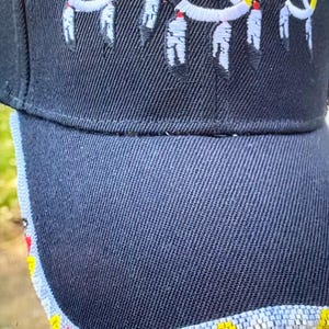 May include: A black baseball cap featuring three embroidered circular medicine wheel designs in red, yellow, black, and white, each with dangling feather motifs. The brim is edged with a band of small, multi-colored beads in a repeating pattern.