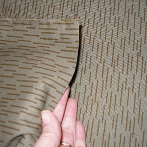 May include: Close-up of a tan fabric with a repeating pattern of brown horizontal and vertical lines. The fabric is being held up, revealing the edge and texture of the material. The pattern gives the fabric a camouflage appearance.