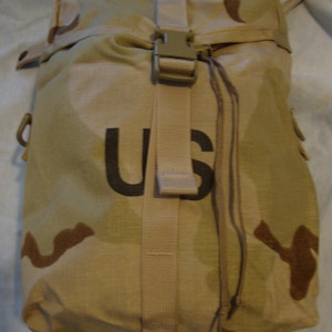 May include: Tan camouflage bag with a drawstring closure and a buckle strap. The bag features the letters "US" in black. The bag is made of a durable fabric and has a military-style design.