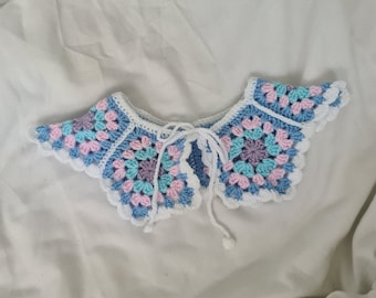 May include: A crocheted collar with a white base and a colourful pattern of blue, pink, and green. The collar has a tie closure at the back.