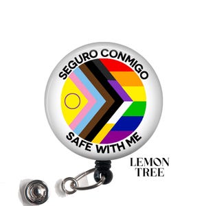 May include: A circular badge reel featuring the Progress Pride flag design with a yellow circle and purple ring. The text SEGURO CONMIGO and SAFE WITH ME is printed in black along the top and bottom edges. A silver metal clip is attached at the base.