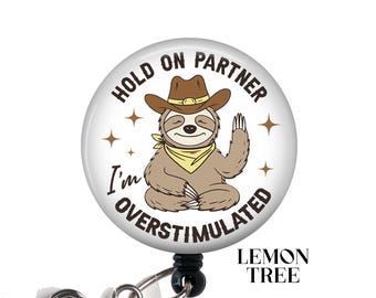 Può includere: Un badge rotondo bianco con un bradipo dei cartoni animati che indossa un cappello da cowboy e medita. Il testo sul badge dice "Hold on partner I'm overstimulated".