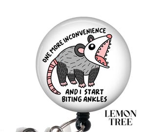 Puede incluir: Insignia redonda blanca con un opossum de dibujos animados con orejas rosas y una boca rosa. El texto en la insignia dice "One more inconvenience and I start biting ankles".