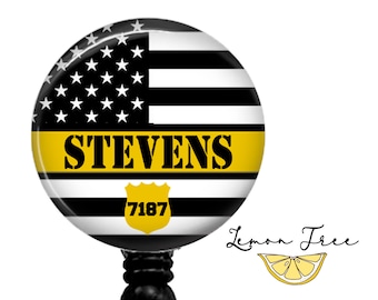 Op de afbeelding: Een ronde badge met een zwart-wit Amerikaans vlagontwerp, een gele streep en de naam "STEVENS" in zwart. Een geel schild met het nummer "7187" staat onderaan. De badge is bevestigd aan een zwarte oprolhaspel.