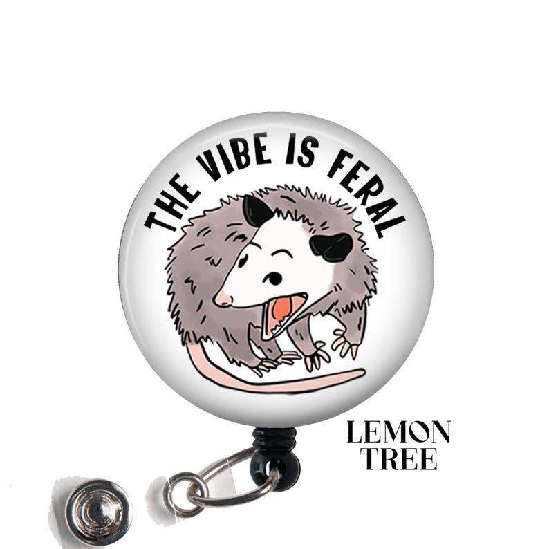 May include: A white button with a black outline and the text "The Vibe Is Feral" in black. A cartoon possum with a white face and black eyes is shown in the center of the button. The possum is facing the right side of the button and has its mouth open. The button is attached to a black retractable badge reel.
