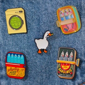 May include: Five enamel pins attached to a blue denim pocket. The pins include a yellow washing machine with green clothes, a white goose with an orange beak, and three rectangular sardine tins with colorful labels and fish illustrations.