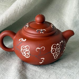 May include: A small, reddish-brown ceramic teapot with a matching lid. The teapot is decorated with white floral and branch designs. It has a rounded body, a curved handle, and a spout. The lid has a small knob on top.